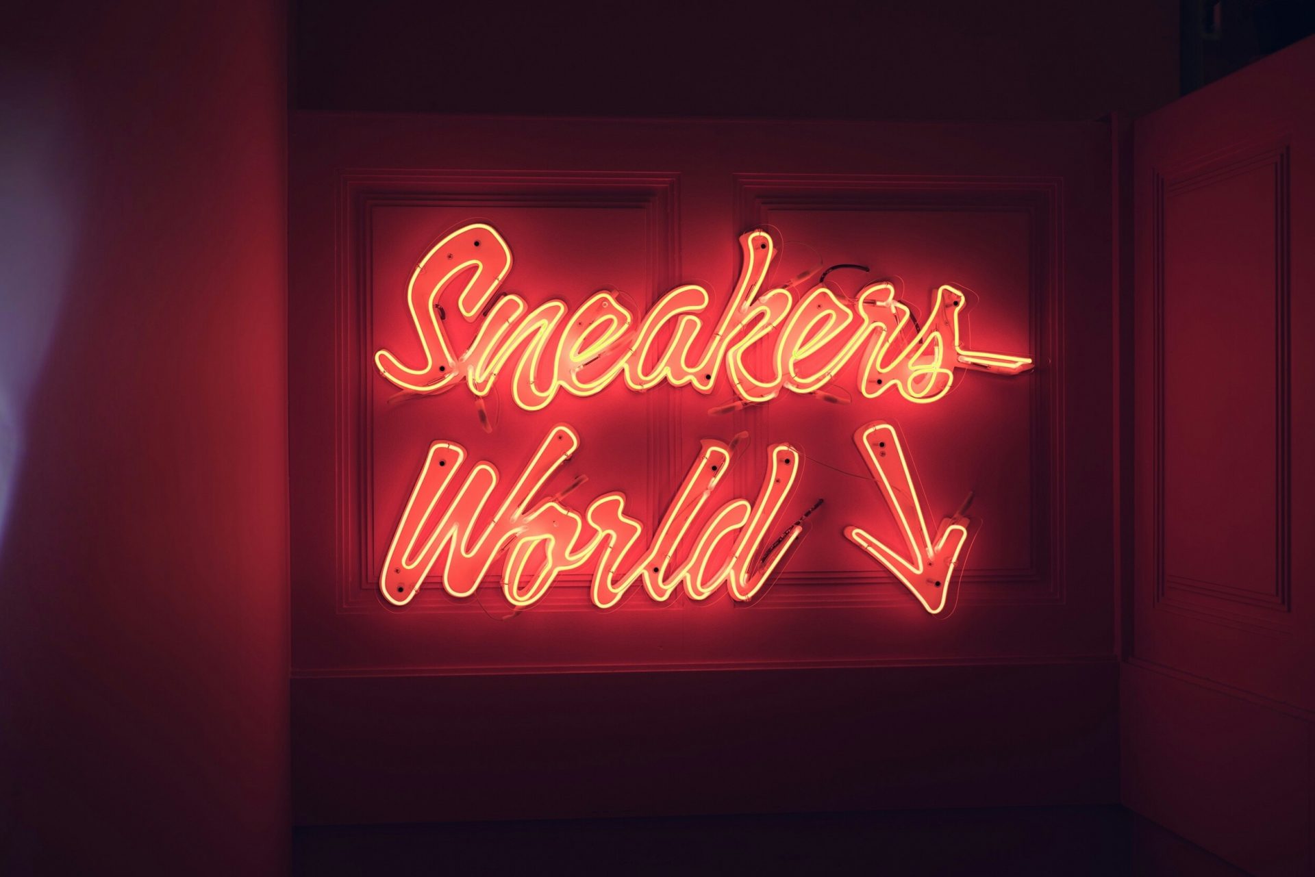 Neon sign reading “Sneakers World” with two downward arrows, glowing red in a dark hallway.